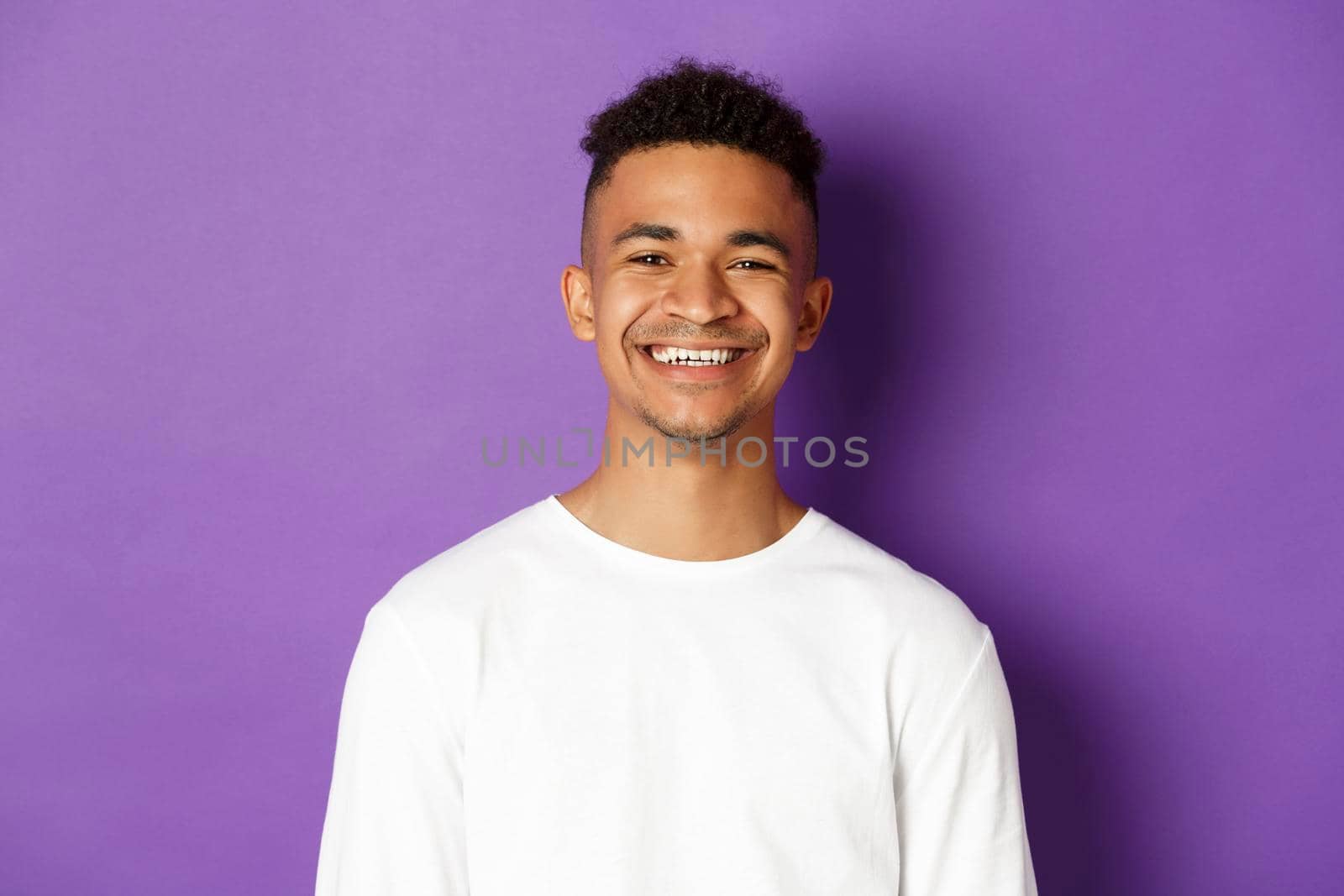 Close-up of young african-american man, smiling pleased, standing satisfied over purple background, wearing white sweatshirt by Benzoix