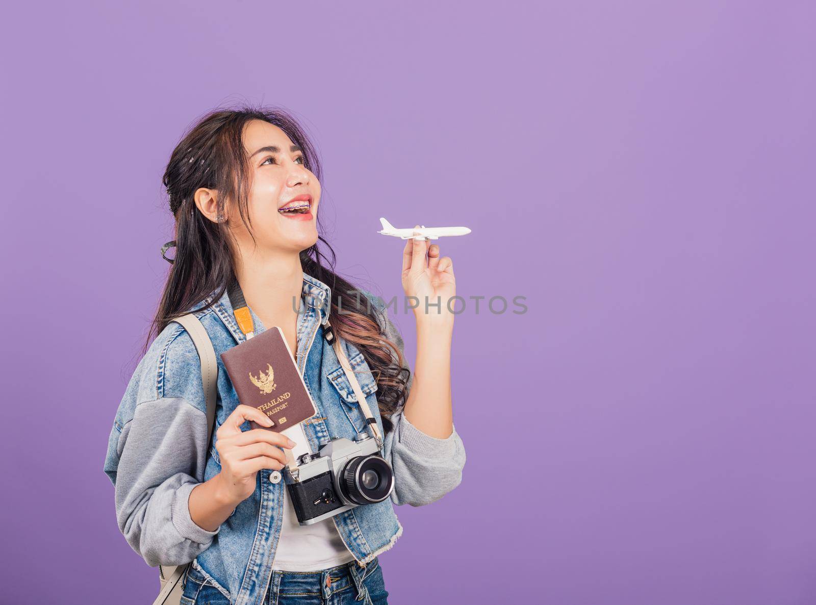 Happy Asian portrait beautiful young woman excited smiling in summer vacation holding airplane toy, passport and vintage photo camera, Thai female ready travel trip isolated on purple background