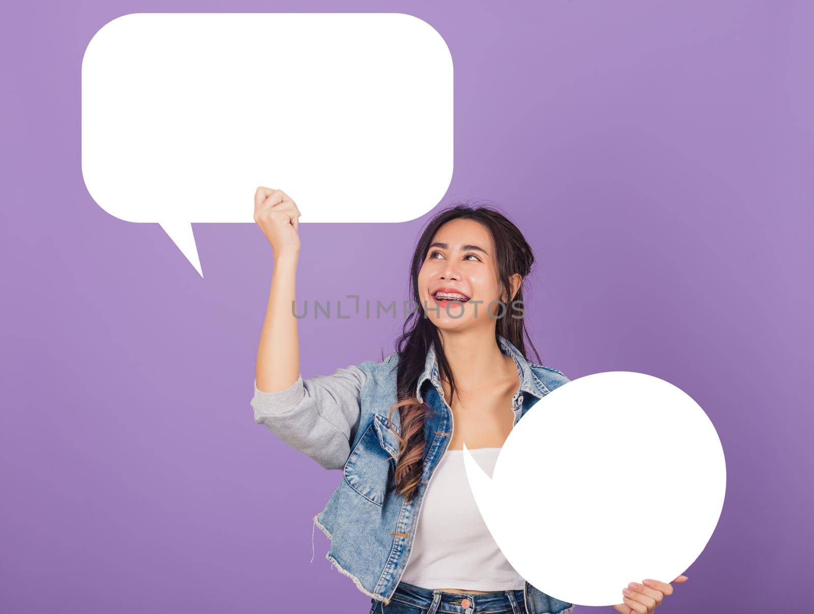 Happy Asian beautiful young woman smiling excited wear denims hold empty speech bubble sign, Portrait female posing show up for your idea looking at bubble, studio shot isolated on purple background