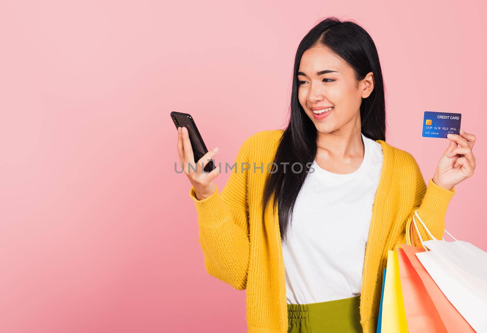 Portrait Asian happy beautiful young woman shopper smiling standing excited holding online shopping bags colorful and credit card for payment on hand in summer, studio shot isolated on pink background