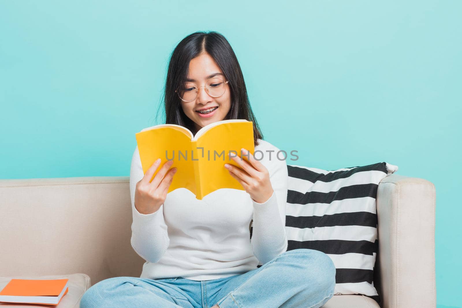 Asian beautiful young woman wearing eyeglasses sitting on sofa reading storybook, portrait relaxation of happy female smiling in living read a book studio shot isolated on blue background