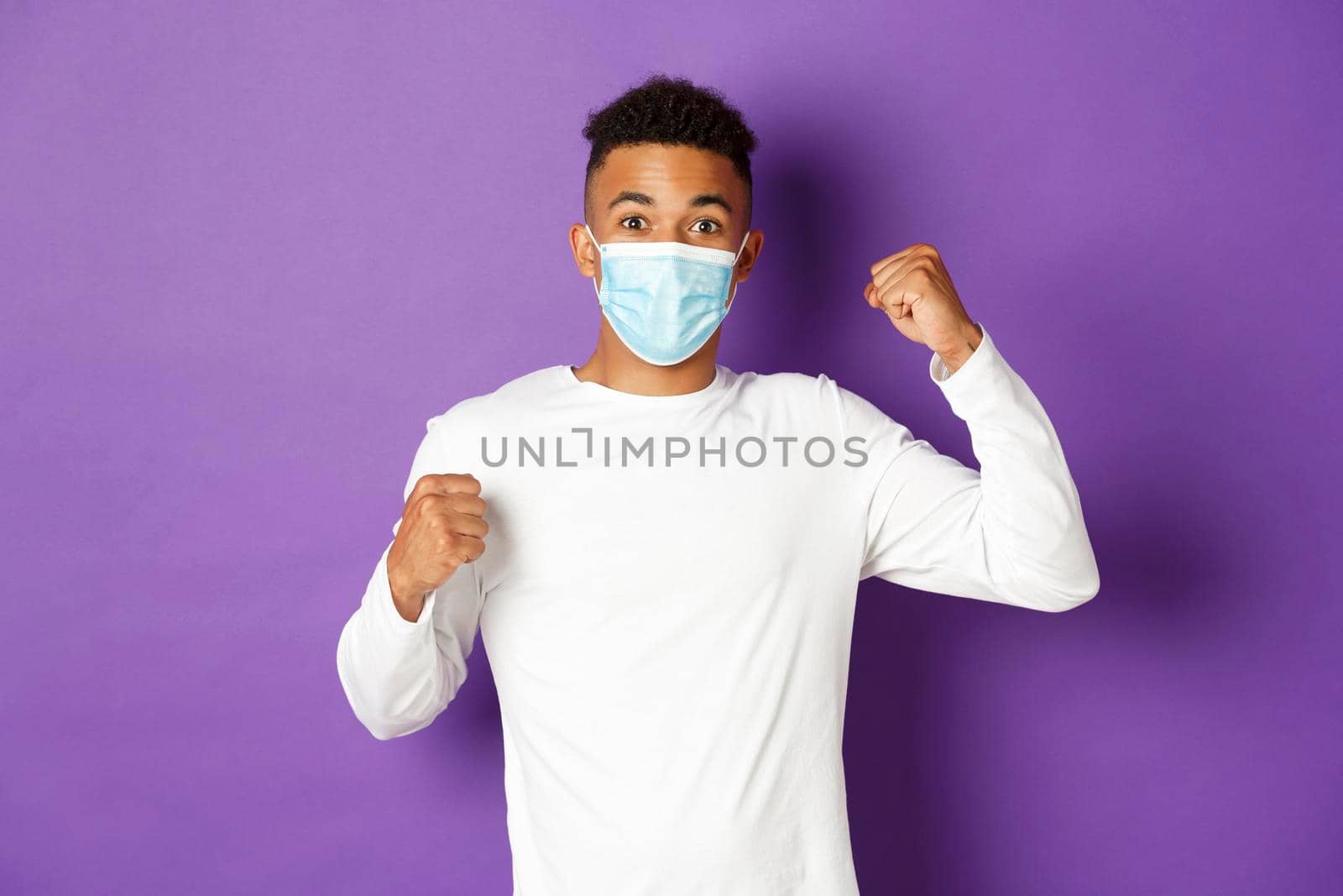 Concept of covid-19, pandemic and social distancing. Young modern african-american man in medical mask, shouting for joy and celebrating something, winning, standing over purple background by Benzoix