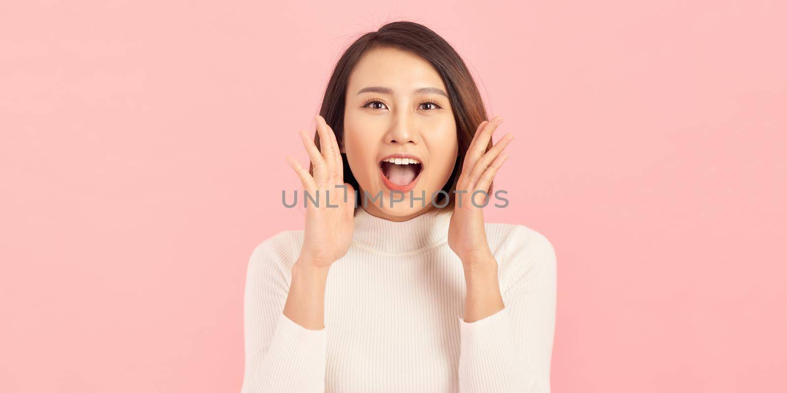 Energetic loud cute beautiful female opening mouth widely, putting hands to mouth, shouting standing isolated over pink background  by makidotvn