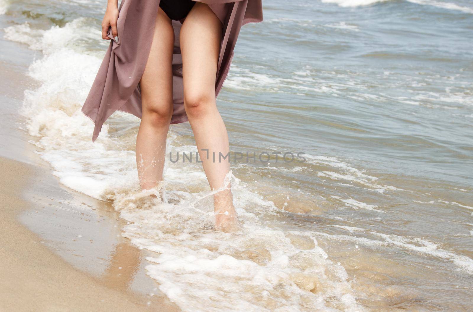 legs of a young woman in black bikini and beige dress standing on sand by sea on sunny summer day
