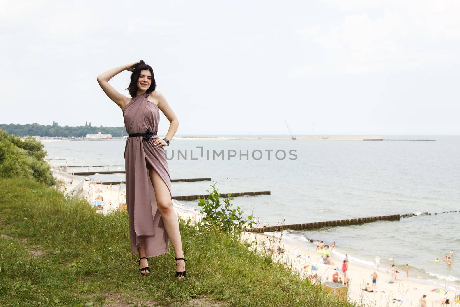 young brunette woman in beige dress standing by the sea by raddnatt