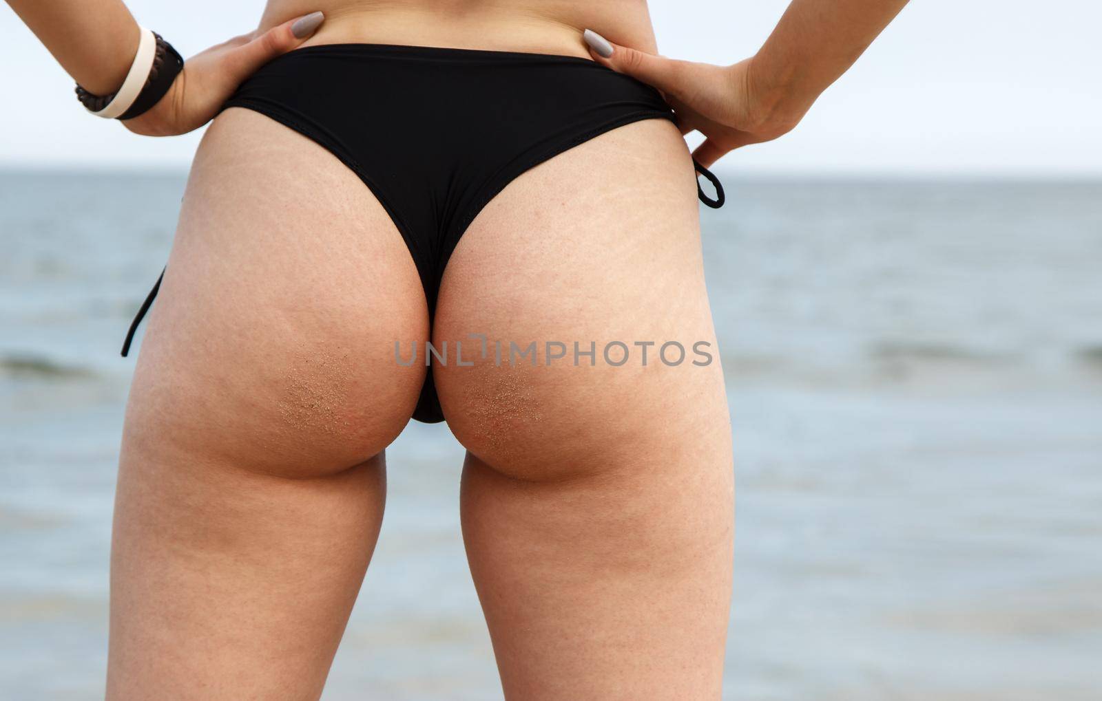 legs of a young woman in black bikini standing on the sand by the sea on sunny summer day. backside closeup