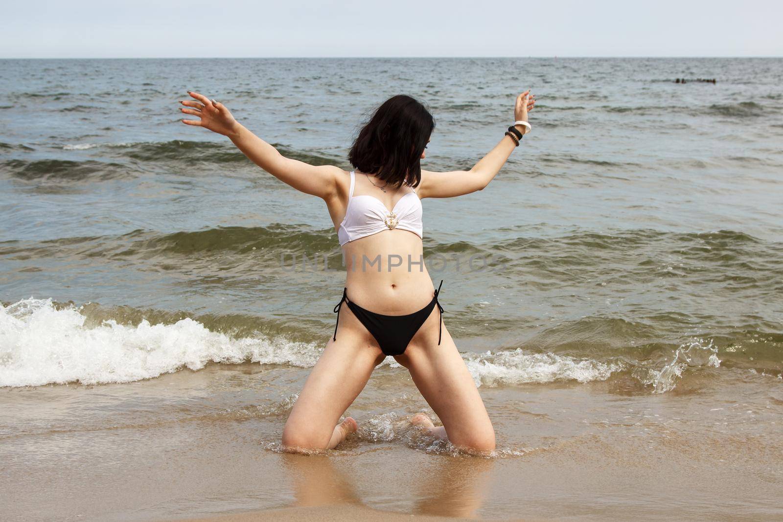 young brunette woman in bikini posing kneeling on the sand by the sea on sunny summer day