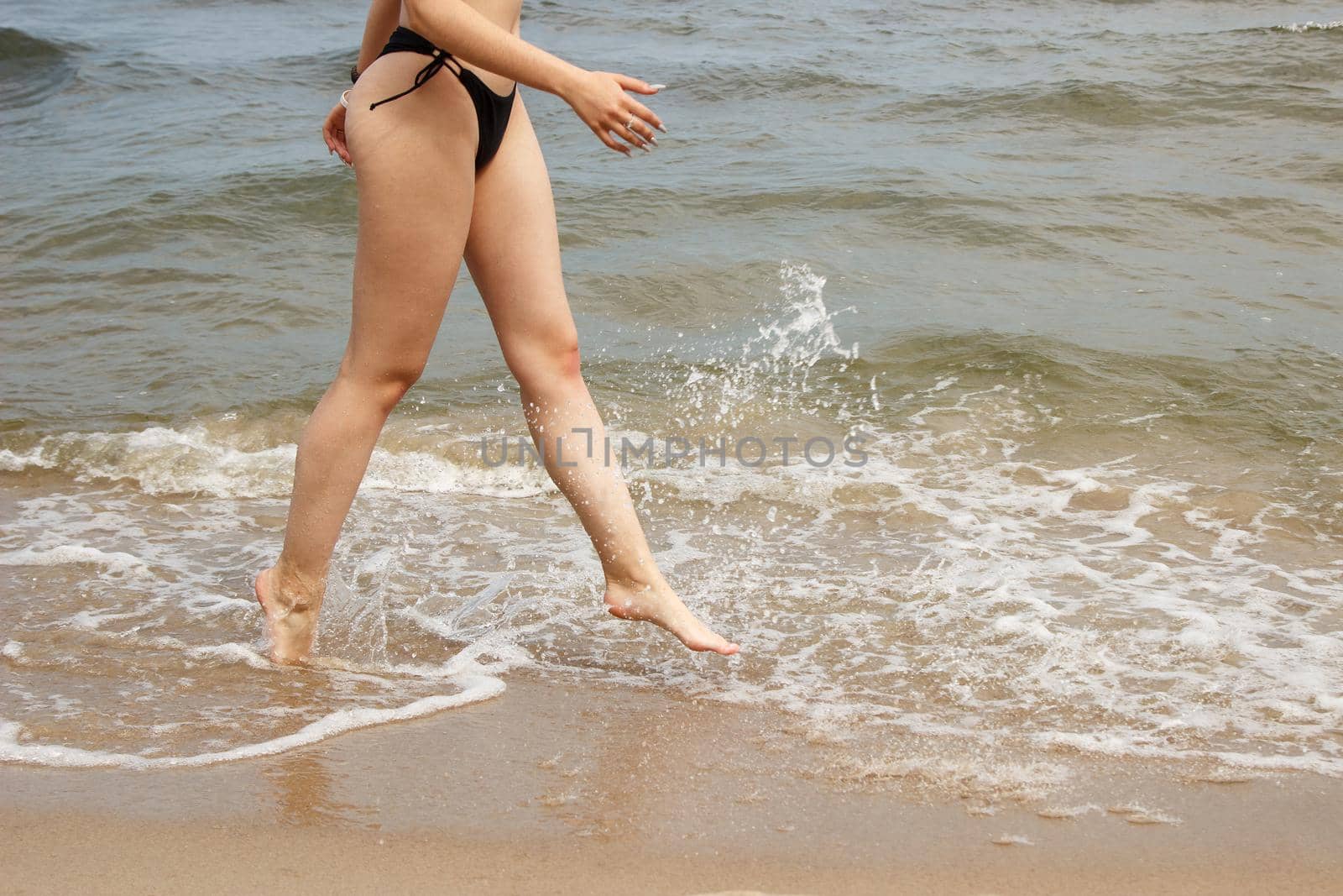 young beautiful girl in a black swimsuit running along the sand of the sea shore on sunny summer day. legs closeup