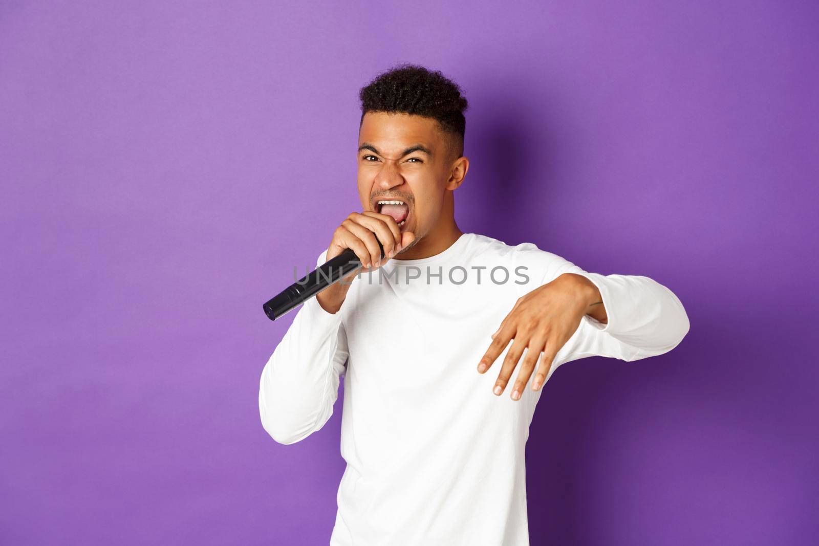 Image of sassy african-american guy singing in microphone, rapping and performing on stage, standing over purple background.