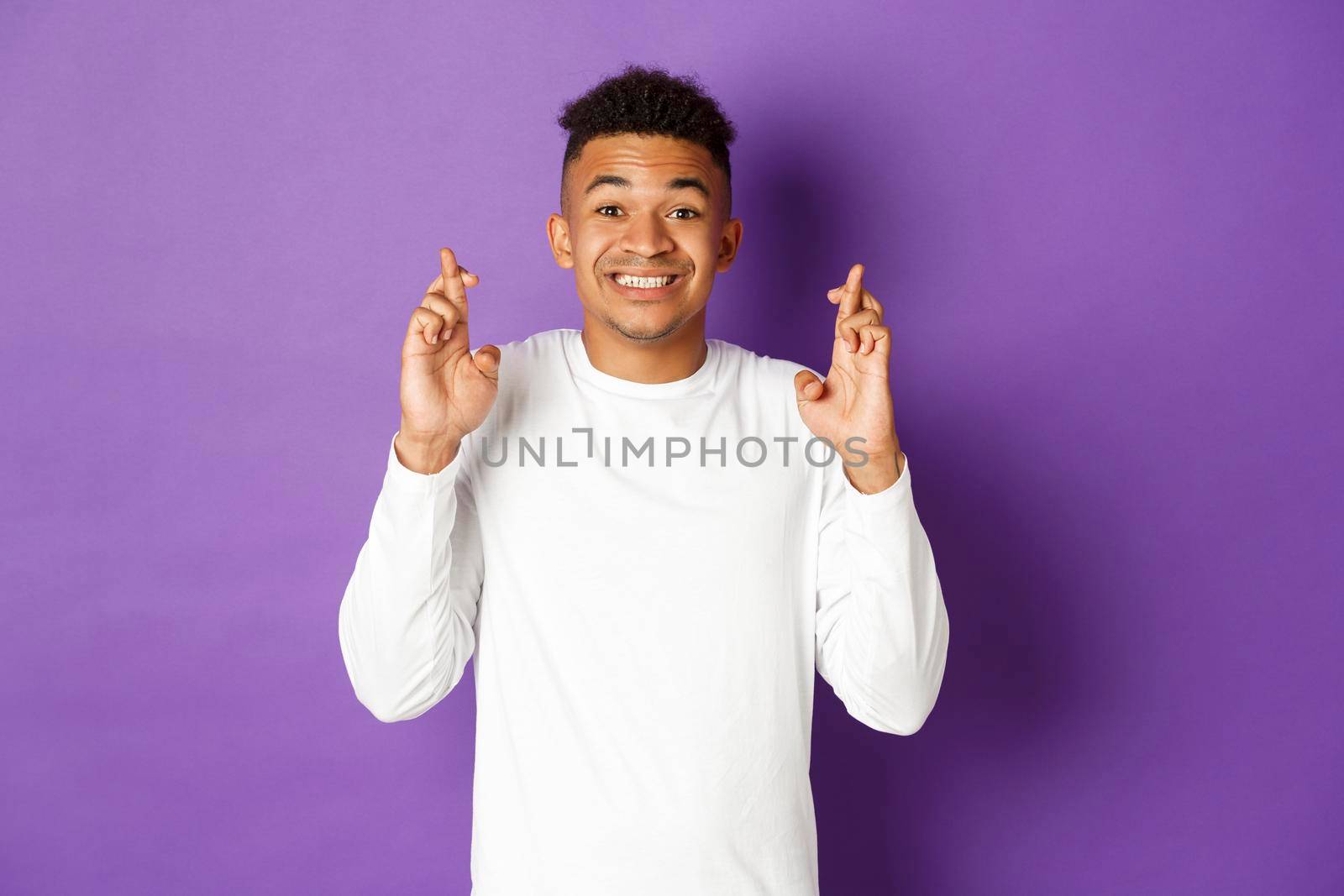 Portrait of hopeful african-american male model in white sweatshirt, cross fingers for good luck and looking at camera, smiling and making wish, standing over purple background by Benzoix