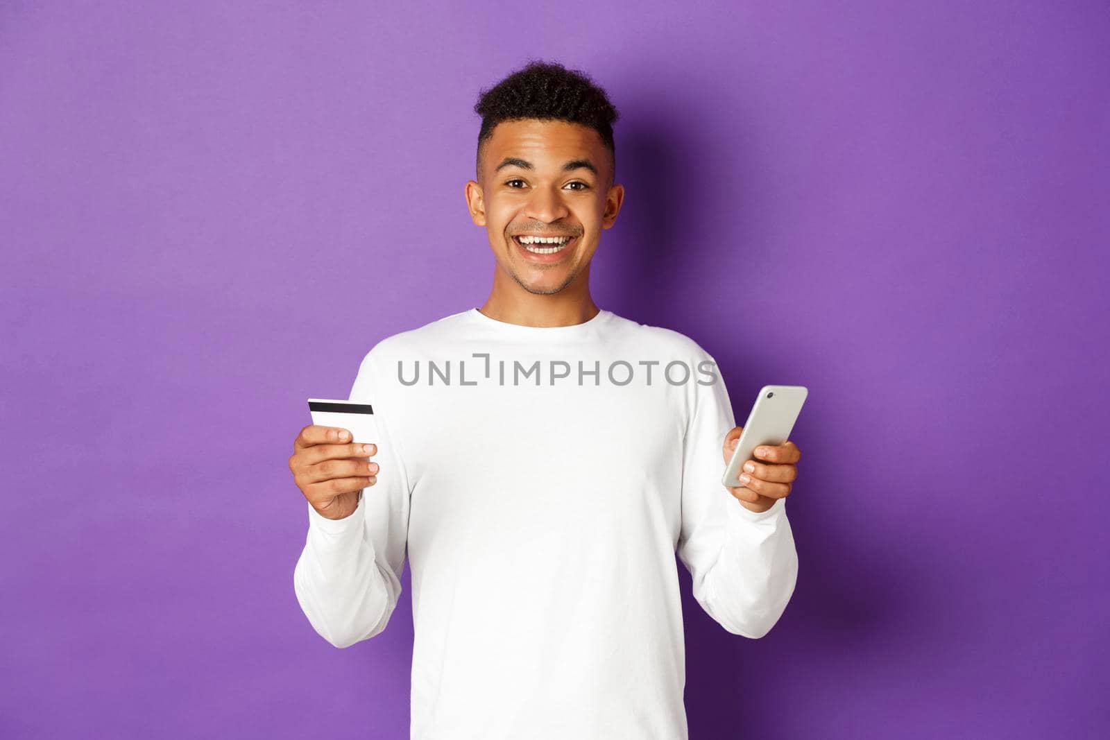 Portrait of handsome african-american man, smiling and looking excited while shopping online, holding credit card and mobile phone, standing over purple background.