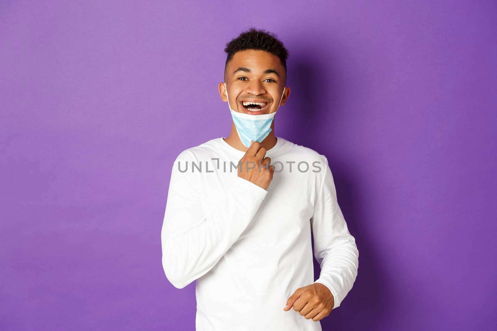 Concept of covid-19, pandemic and social distancing. Happy african-american man taking-off medical mask and breathing freely, standing over purple background.