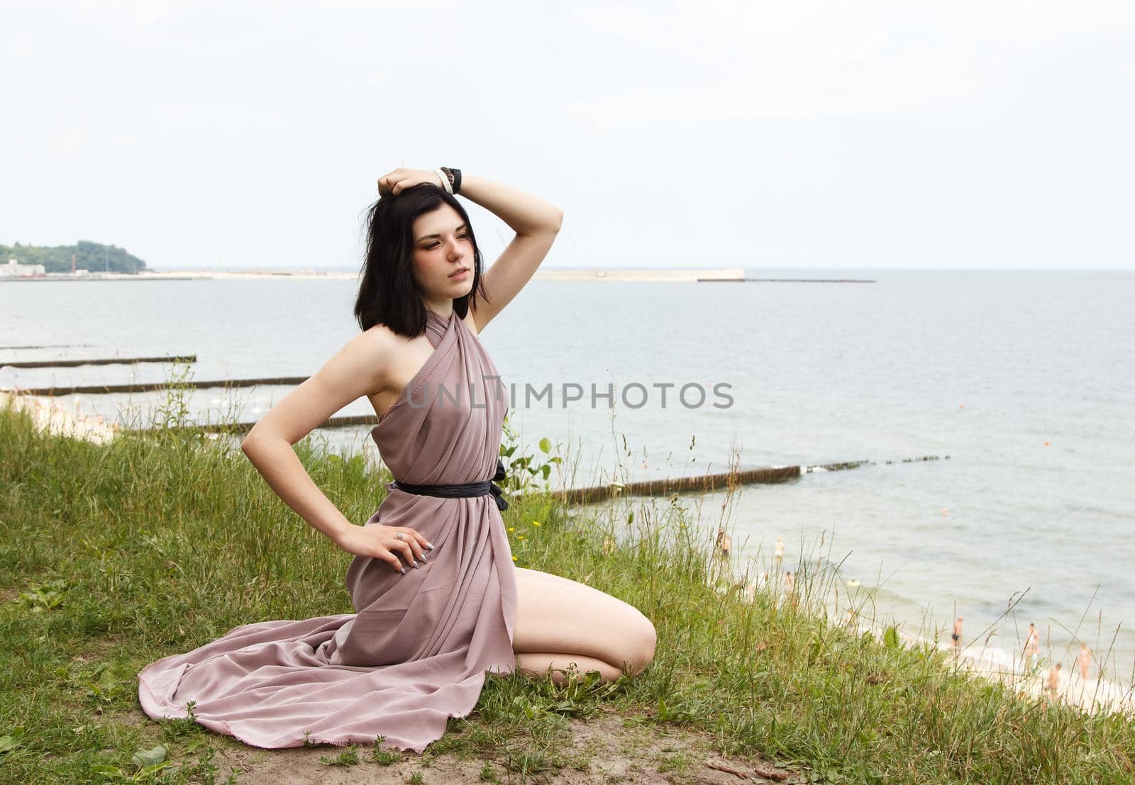 young brunette woman in beige dress sitting by the sea on summer day