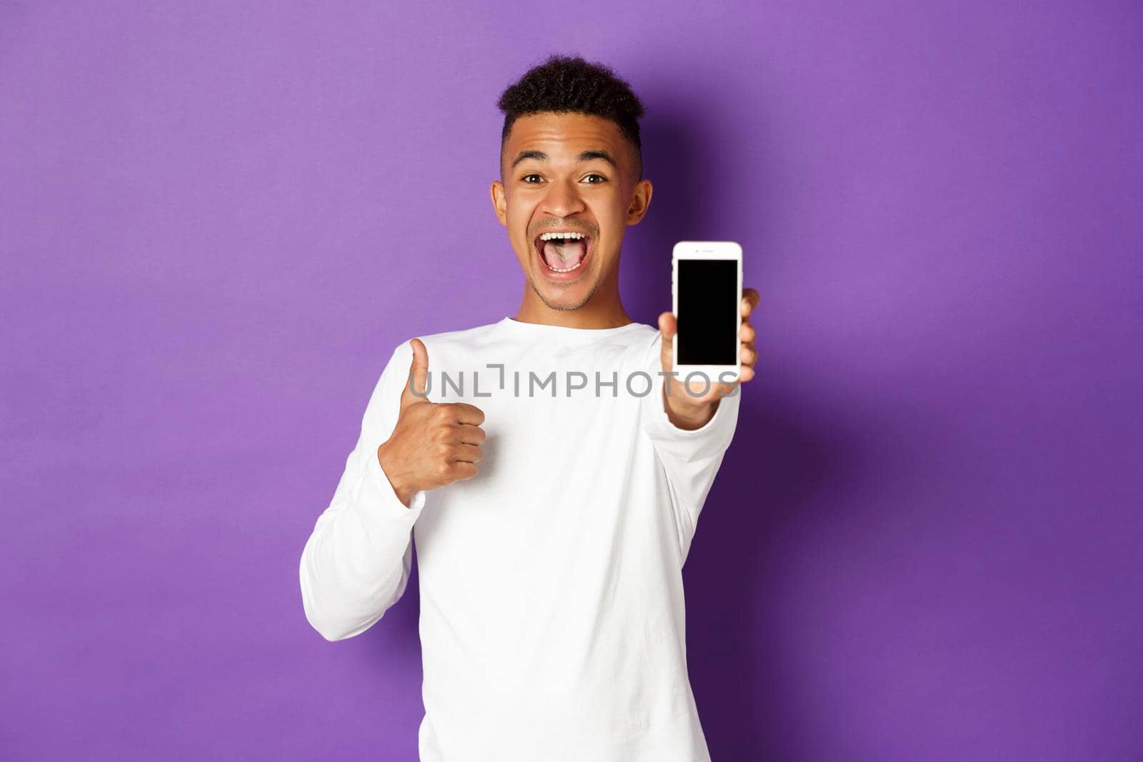 Image of african-american guy in white sweatshirt, smiling and showing thumbs-up in approval, showing mobile phone screen, standing over purple background.