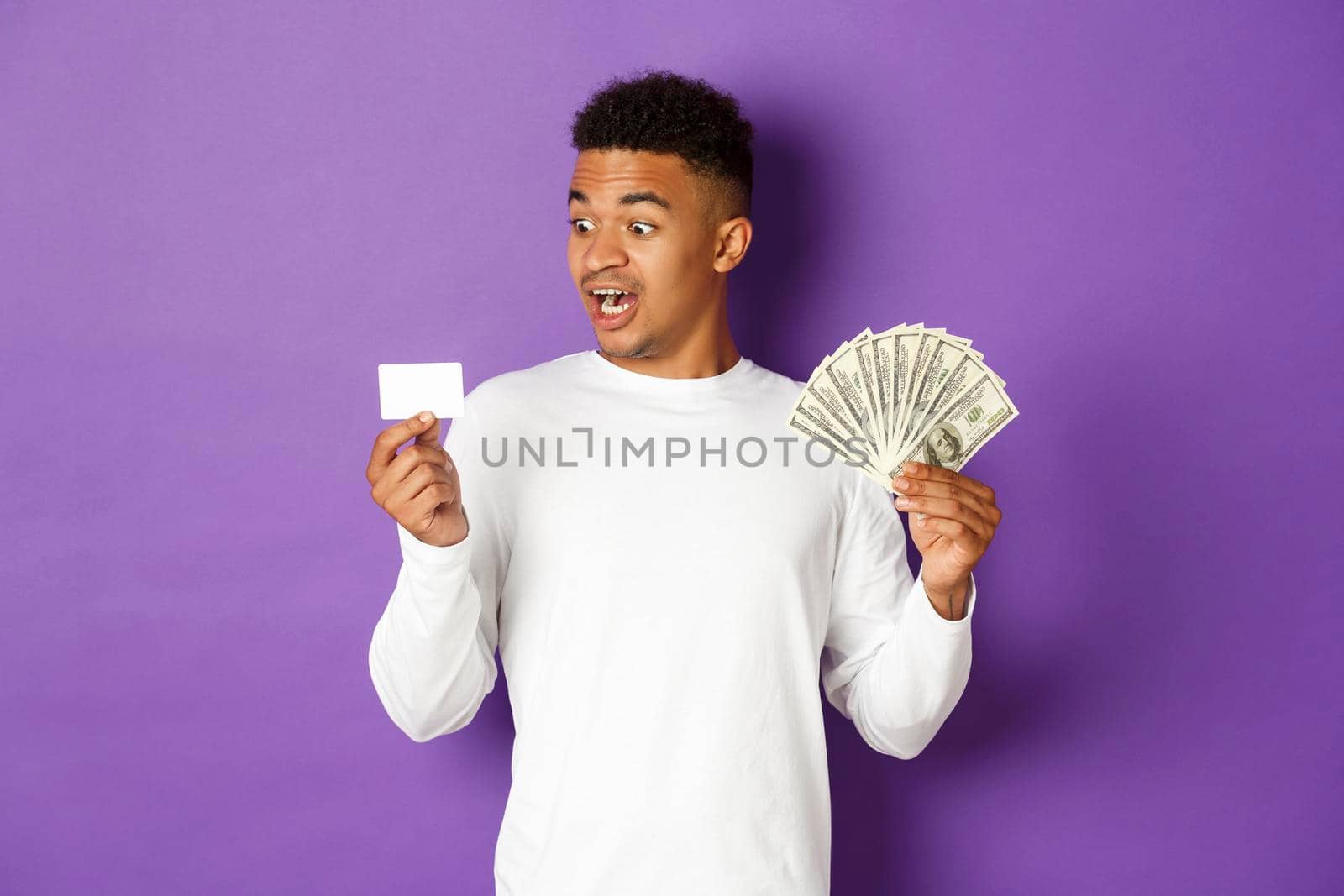 Image of handsome african american guy, showing money and looking excited at credit card, standing over purple background.