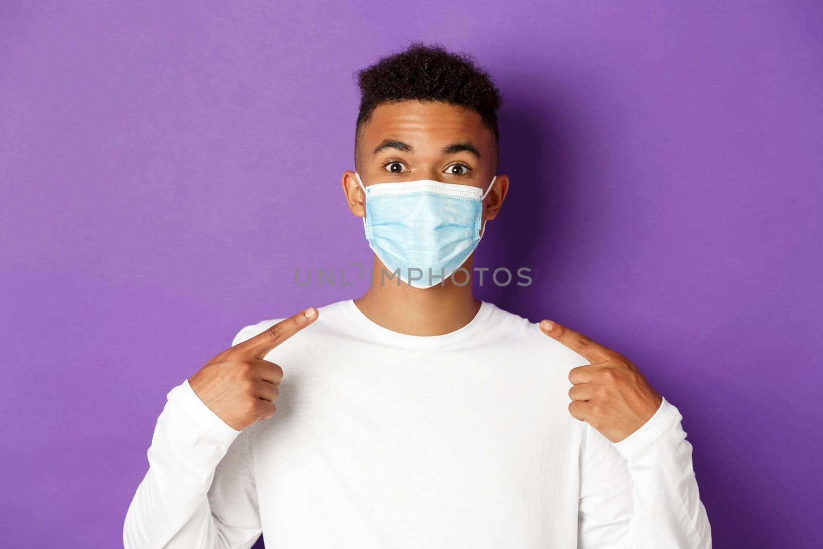 Concept of coronavirus, quarantine and social distancing. Close-up of young african-american man smiling, recommending to wear medical mask during covid-19, standing over purple background.