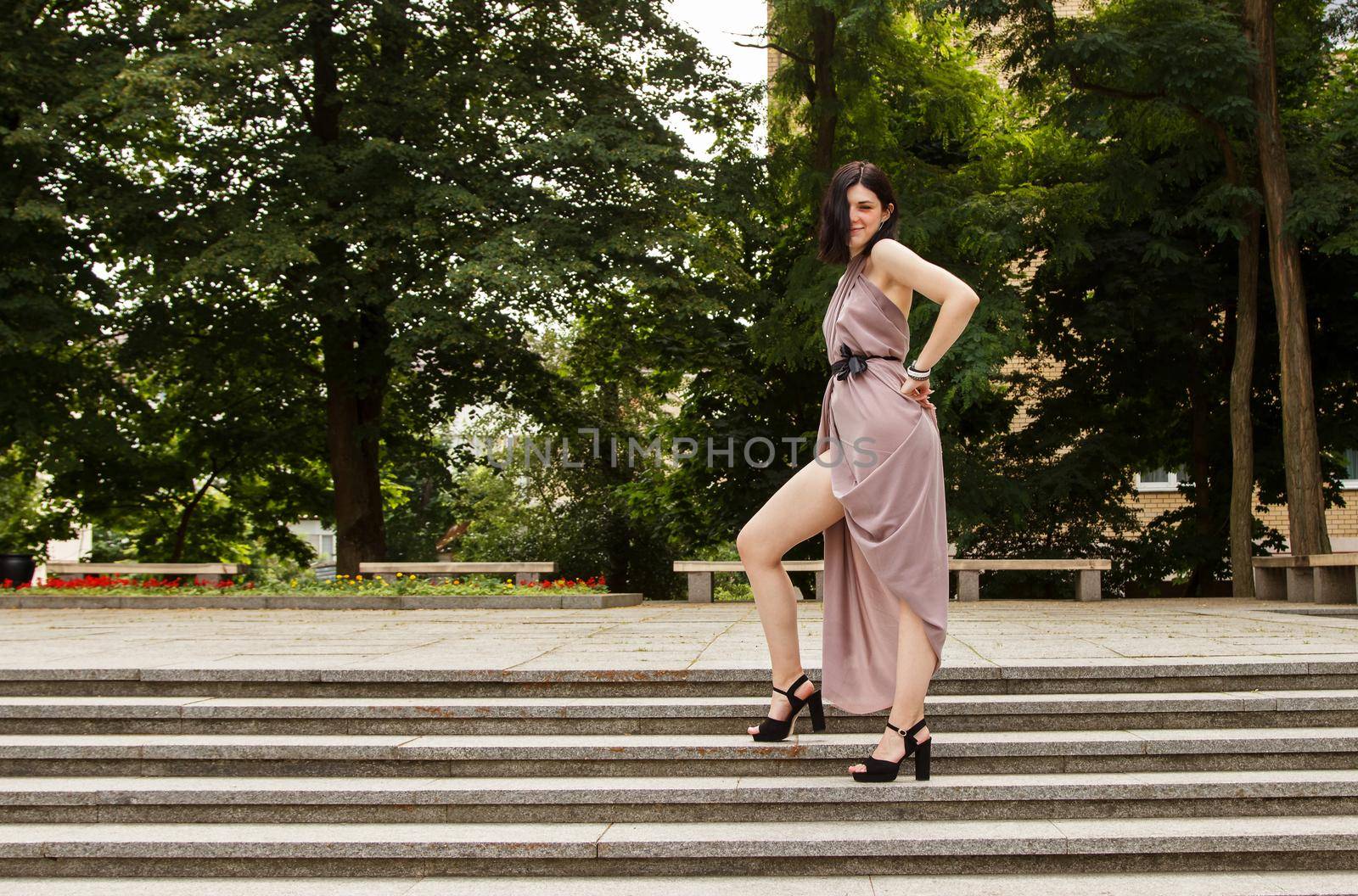 young beautiful brunette woman in beige dress standing on the stairs in the city park on sunny summer day
