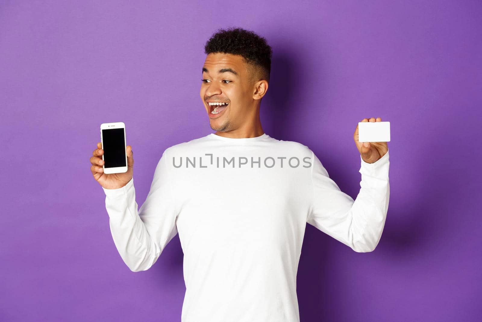 Portrait of handsome african-american man, looking amazed at mobile phone screen and showing credit card, standing over purple background by Benzoix