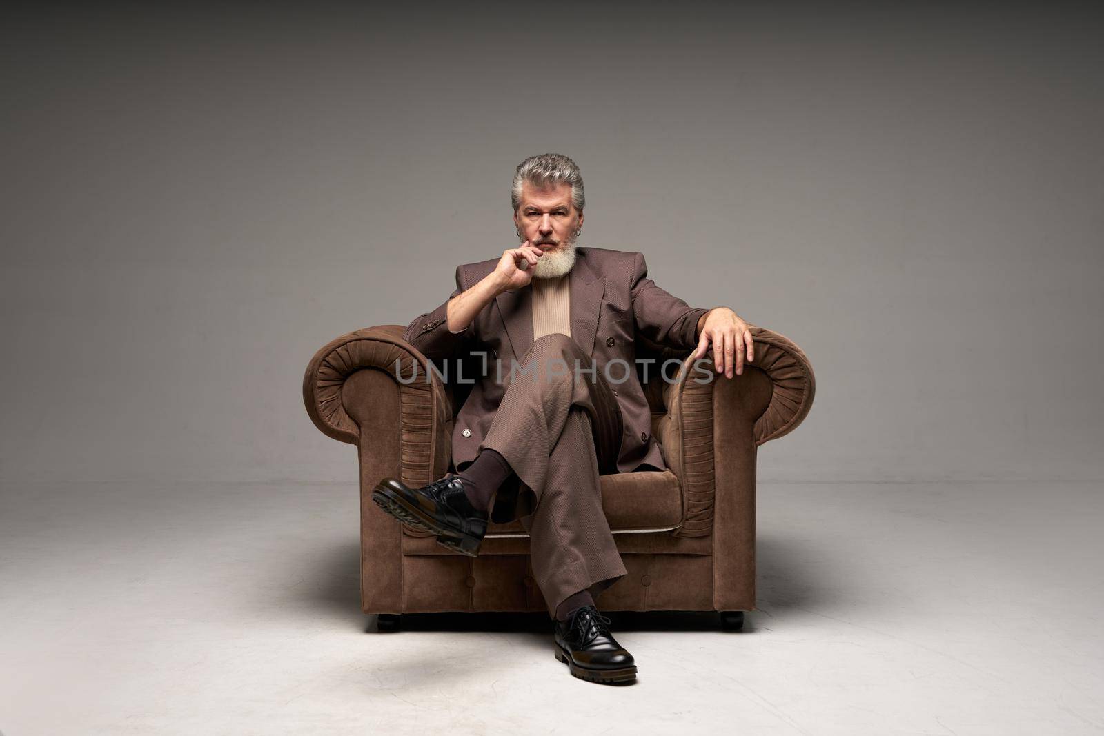 Full length shot of pensive mature businessman with beard wearing elegant suit looking at camera, sitting in armchair while posing isolated over light gray background by friendsstock