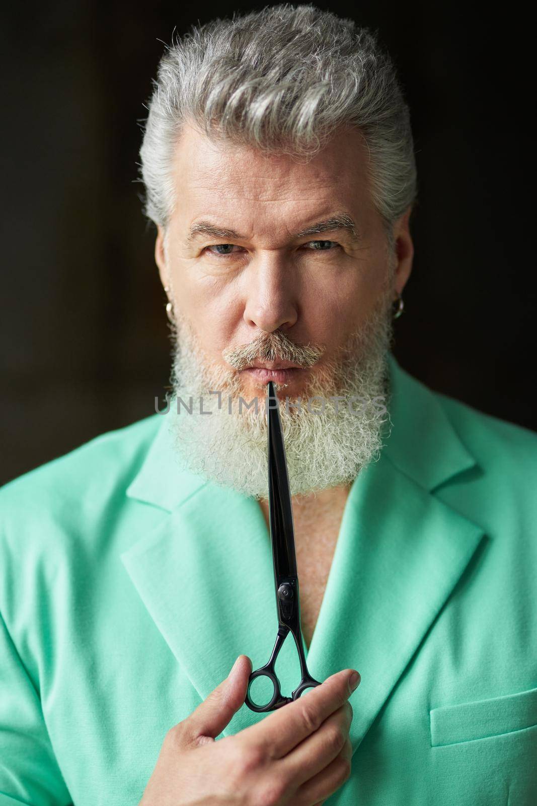 Portrait of serious gray haired mature man with beard wearing colorful outfit looking at camera, holding sharp barber scissors near his face, posing over dark background. Professional occupation