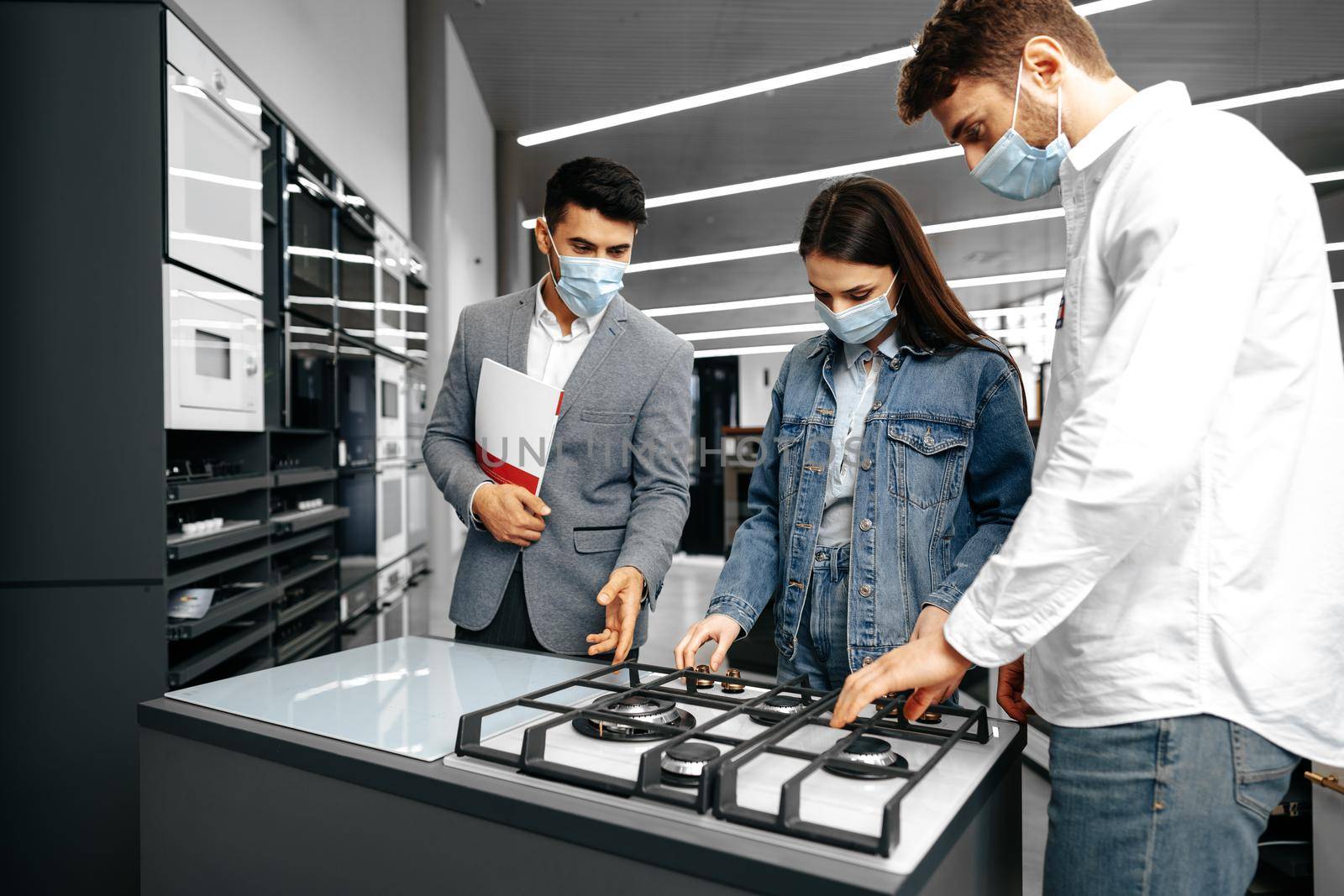 Shop assistant in hypermarket shows new model of gas stove to young couple, all wearing medical masks by Fabrikasimf