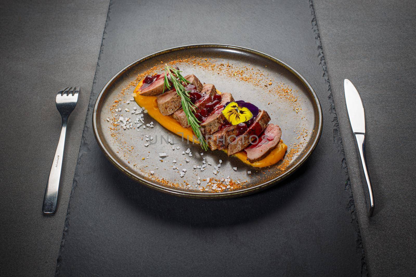 Glazed pork fillet with pumpkin puree and cranberry sauce on a dark plate. Sliced pork tenderloin or sirloin decorated with rosmarin and edible flowers.
