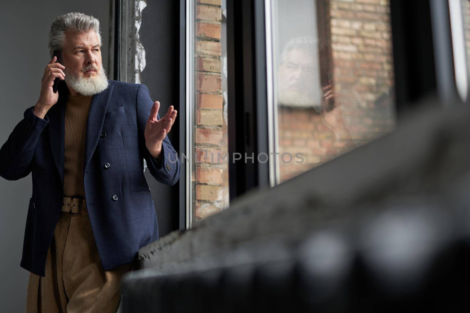 Handsome mature man in business casual wear looking out a large window, making a call using smartphone while standing in loft interior. Lifestyle, people concept