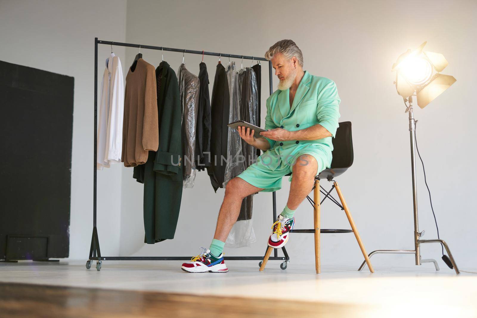 Full length shot of stylish middle aged man wearing colorful outfit using tablet pc while sitting next to clothes rail and studio spotlight over white background. Fashion photoshoot, style concept