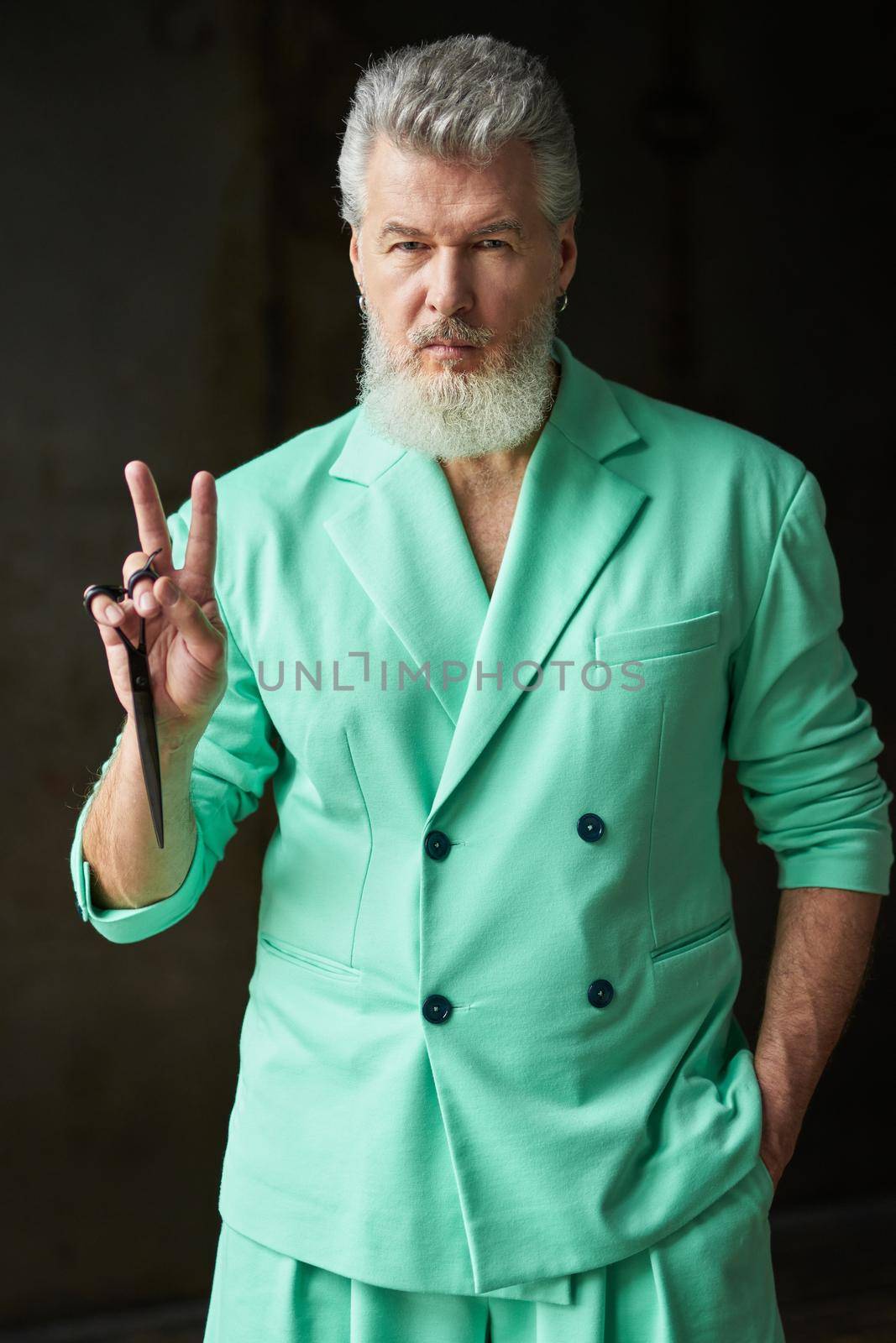 Cool gray haired middle aged man with beard wearing colorful outfit looking at camera, showing peace sign while holding sharp barber scissors, posing over dark background. Professional occupation