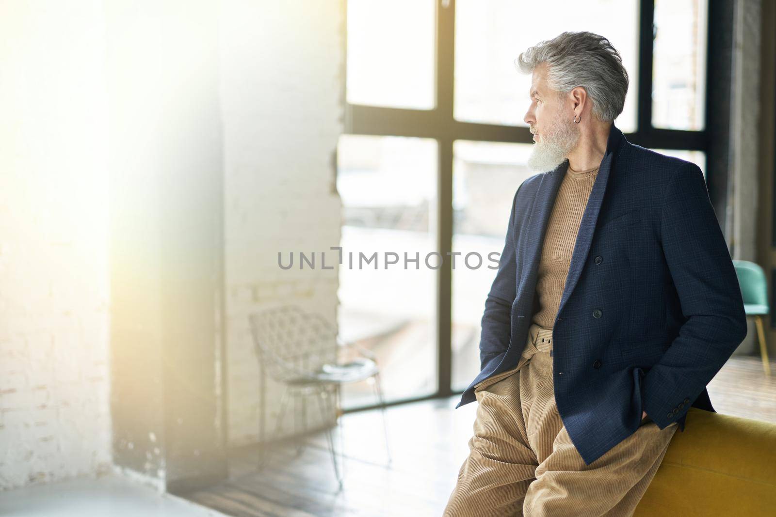 Stylish confident mature man looking aside while posing, leaning on sofa indoors by friendsstock