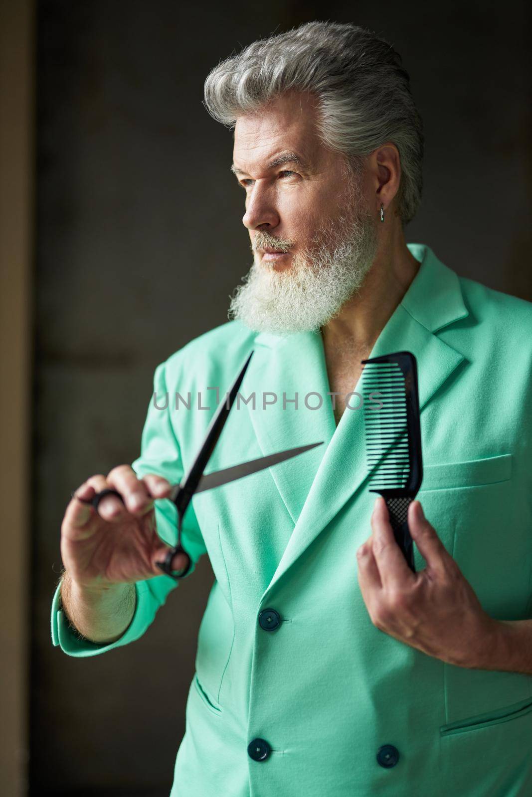 Portrait of classy gray haired mature man with beard wearing colorful outfit looking away, holding sharp barber scissors and hair comb, standing over dark background. Professional occupation concept