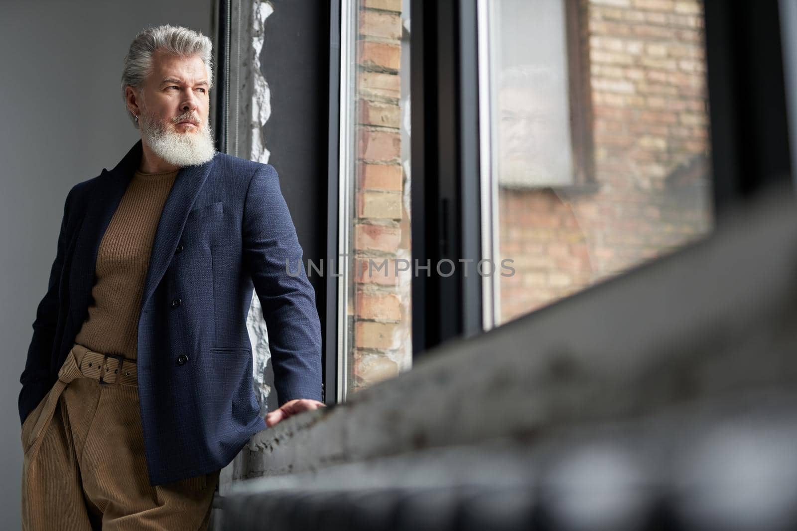 Pensive middle aged man in business casual wear looking out a large window, being thoughtful while standing in loft interior. Lifestyle, people concept