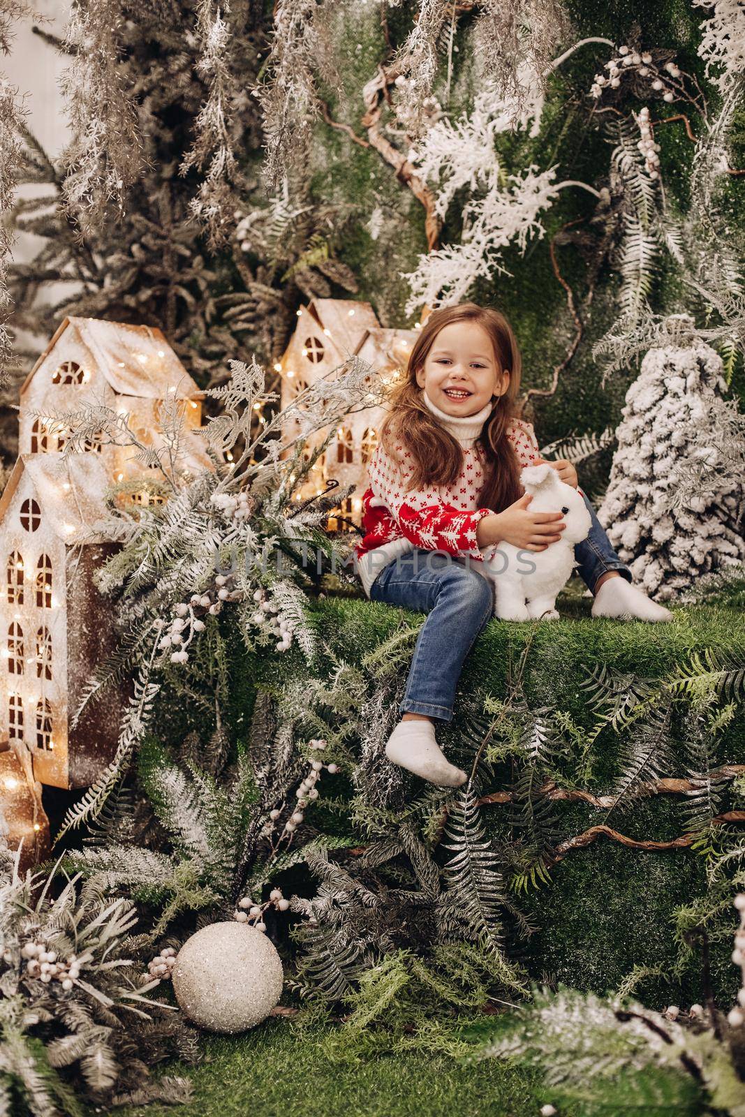 Stock photo of lovely smiling brunette girl in red and white sweater and jeans sitting in Christmas decorations with white toy rabbit, looking at camera.