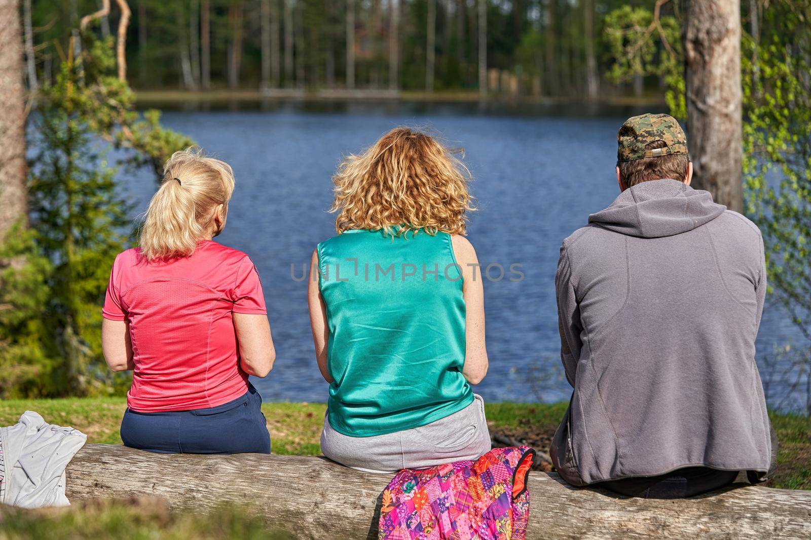 A man and two women sit on the shore of a forest lake with their backs to the camera. People admire the lake