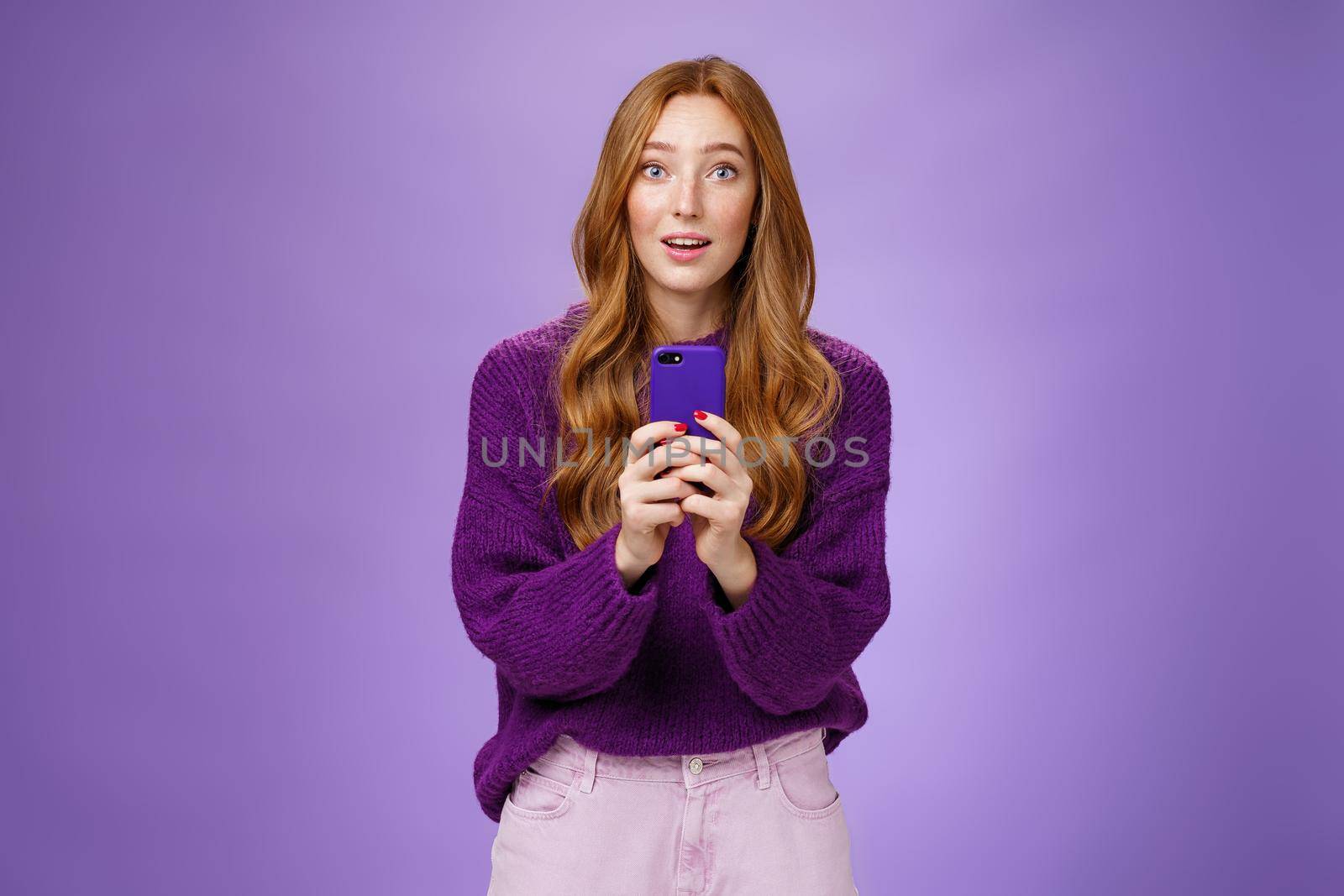 Impressed speechless cute redhead female actor receiving awesome surprising message on mobile phone open mouth from amazement and raising eyebrows astonished, posing over purple wall. Technology and people emotions concept