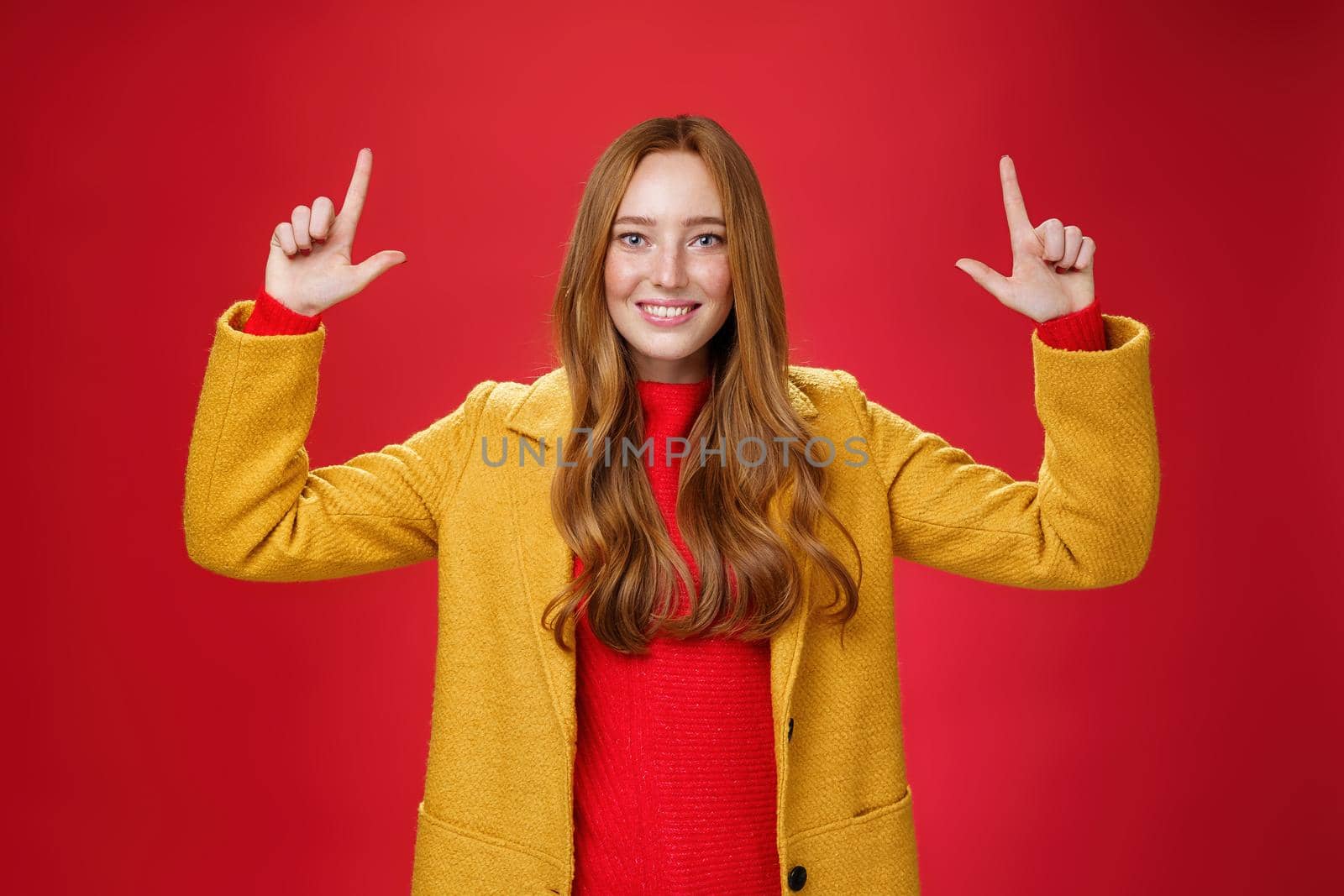 Indoor shot of redhead attractive woman in yellow fall coat raising hands promoting advertisement as pointing up and smiling broadly with satisfied pleasant expression over red background by Benzoix