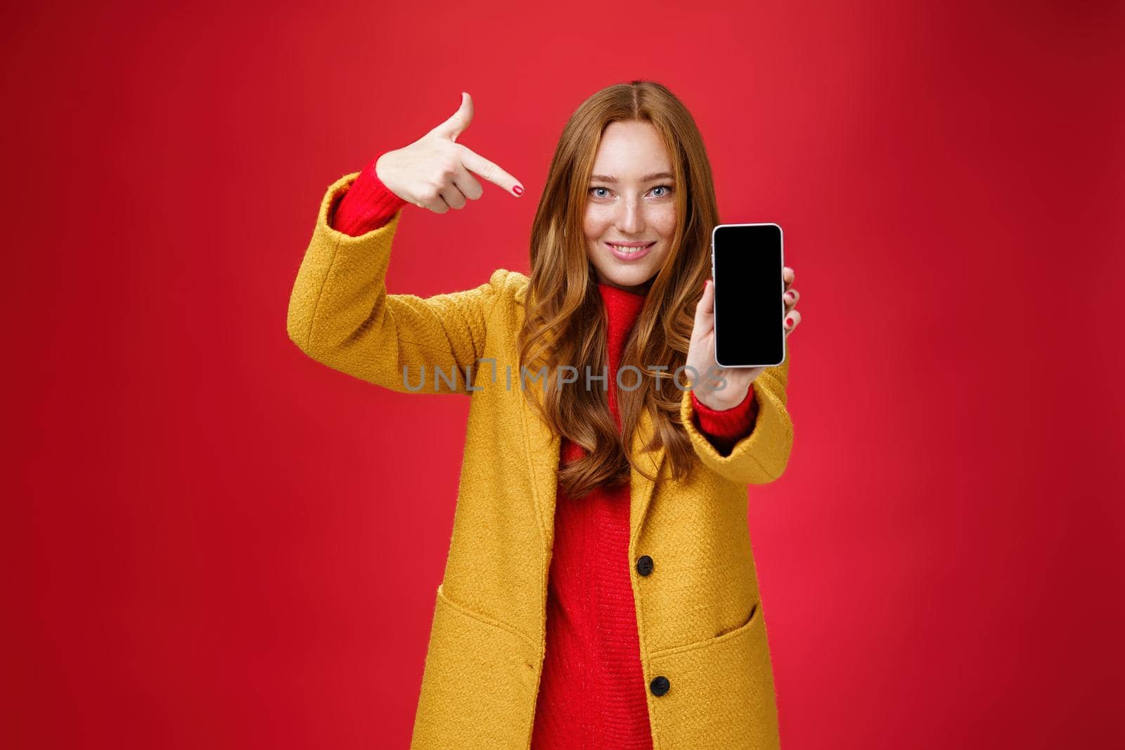 Pick this phone you never regret. Portrait of friendly-looking attractive and confident redhead glamour girlfriend in yellow coat showing smartphone pointing at mobile as smiling broadly at camera.
