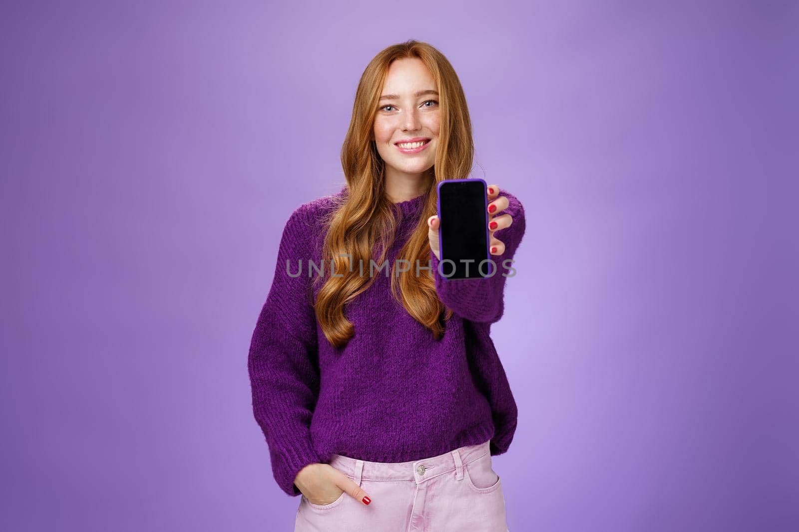 Girl shows smartphone screen at camera to ask opinion of friend smiling broadly with optimistic and joyful expression holding hand in pocket promoting mobile phone or app over purple background by Benzoix