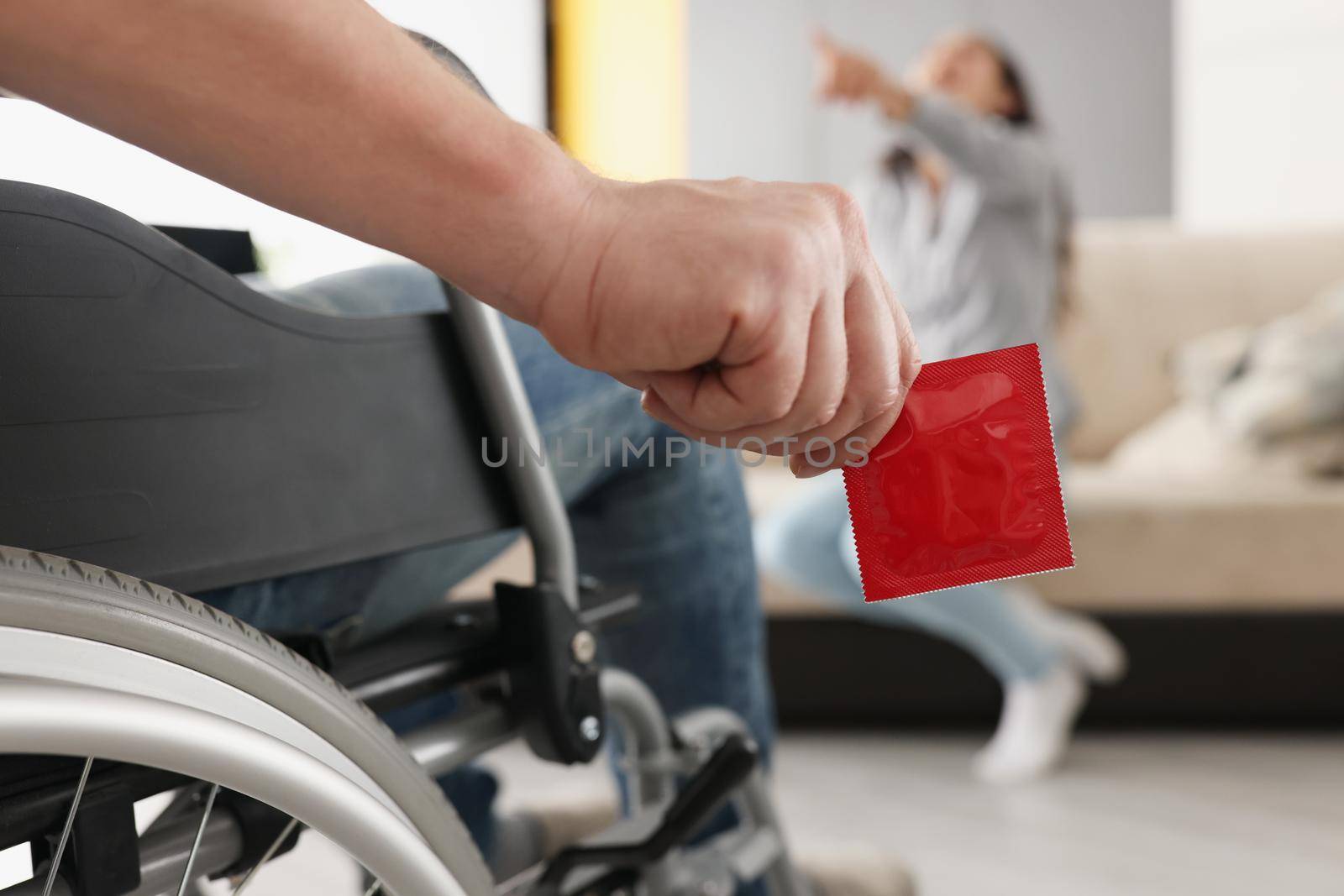 Close-up of man sit in wheelchair hold package of condom, woman laugh on couch pointing with finger. Disabled people, intimate life with disability concept
