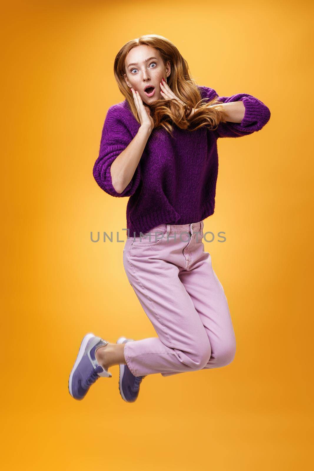 Impressed and surprised amazed good-looking redhead female in purple sweater open mouth astonished and holding hands on cheeks as jumping popping eyes amused over orange background.