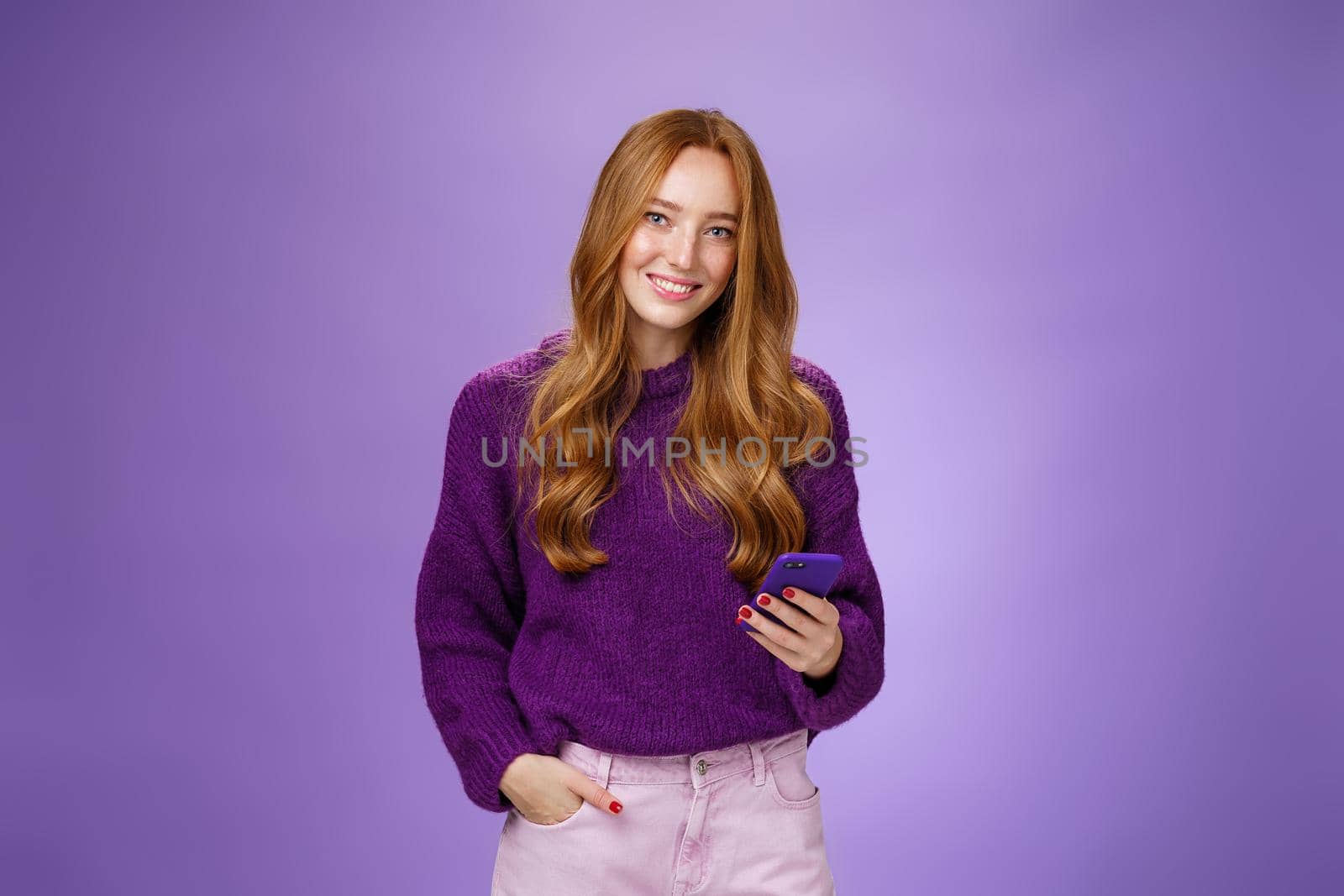 Friendly-looking relaxed and joyful nice 20s female student writing post in social network using mobile phone holding one hand in pocket and texting friend with smartphone, smiling satisfied by Benzoix