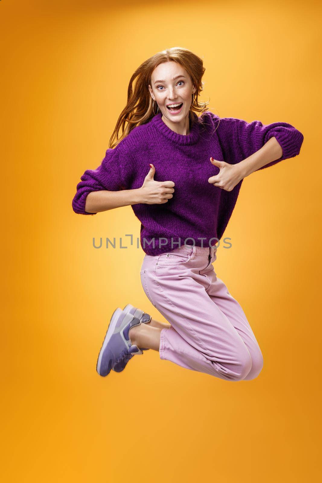 Excited and happy funny young redhead female in purple sweater jumping from happiness and satisfacion showing thumbs up gesture in approval giving positive reply and liking awesome clothes by Benzoix