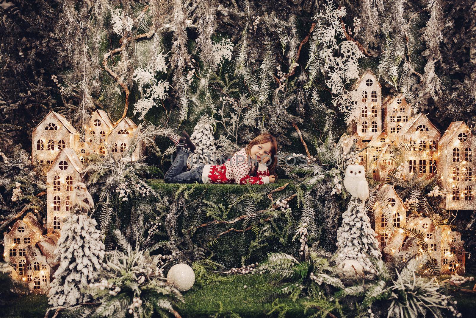 Beautiful brunette female kid in winter sweater and jeans laying on green lawn surrounded by illuminated handmade houses and fir branches covered with artificial snow. Christmas fairytale.