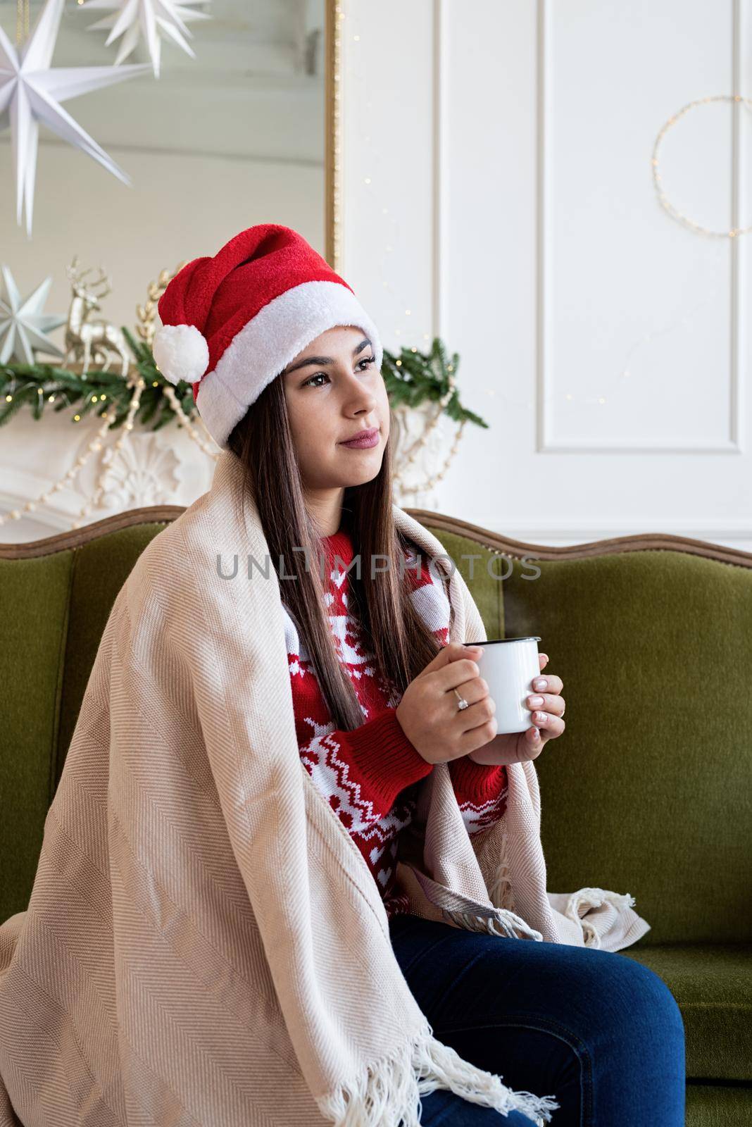 Merry Christmas and Happy New Year. Young brunette woman in santa hat and cozy plaid sitting on green couch alone in a decorated for Christmas living room, having cup of hot drink