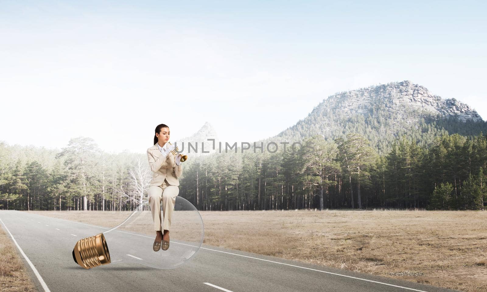 Attractive woman playing trumpet brass on asphalt road. Young businesslady in white business suit and gloves sitting on big light bulb with music instrument. Musician practicing and performing outdoor
