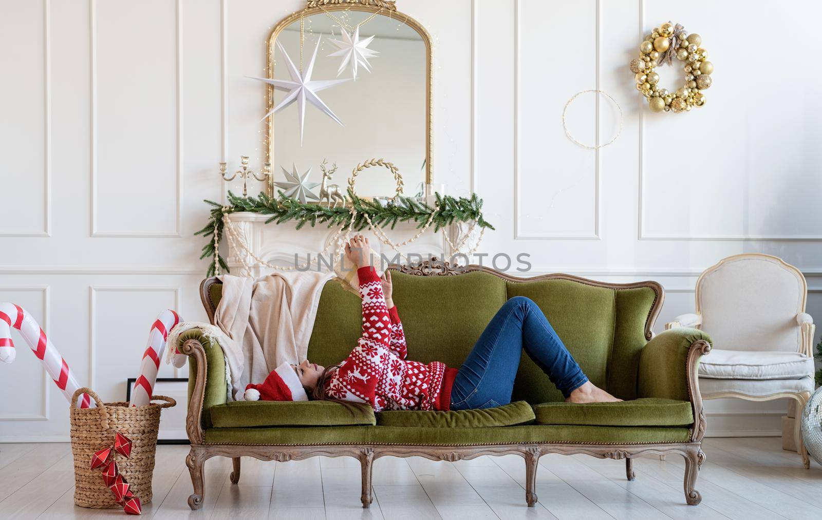 Merry Christmas and Happy New Year. Young brunette woman in santa hat lying on green couch alone in a decorated for Christmas living room