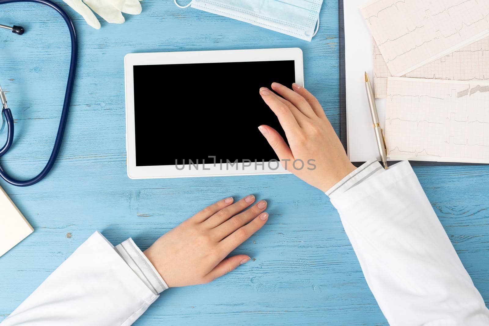 Top view of doctor hands with tablet computer. Cardiac examination in hospital. Therapist sitting at blue wooden desk with stethoscope, medical gloves and mask. Examination and consultation in clinic