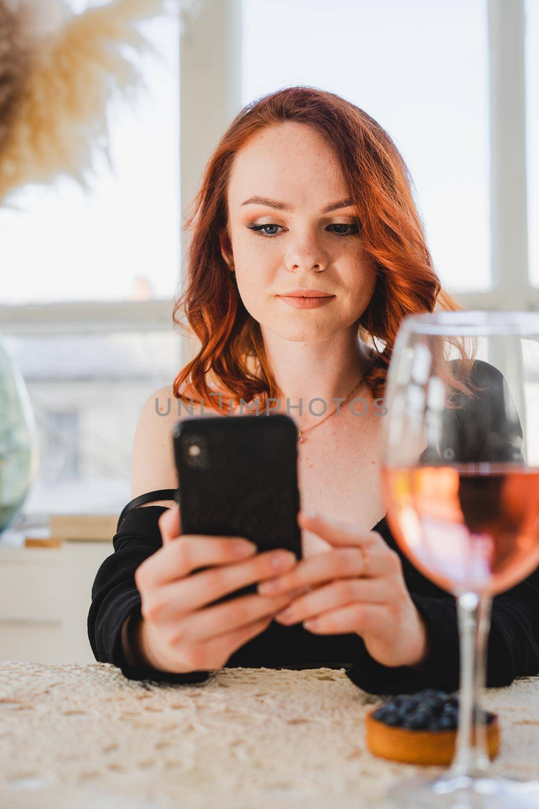 Beautiful girl with red hair holding a smartphone. There's a glass of rose wine on the table. The girl is resting.
