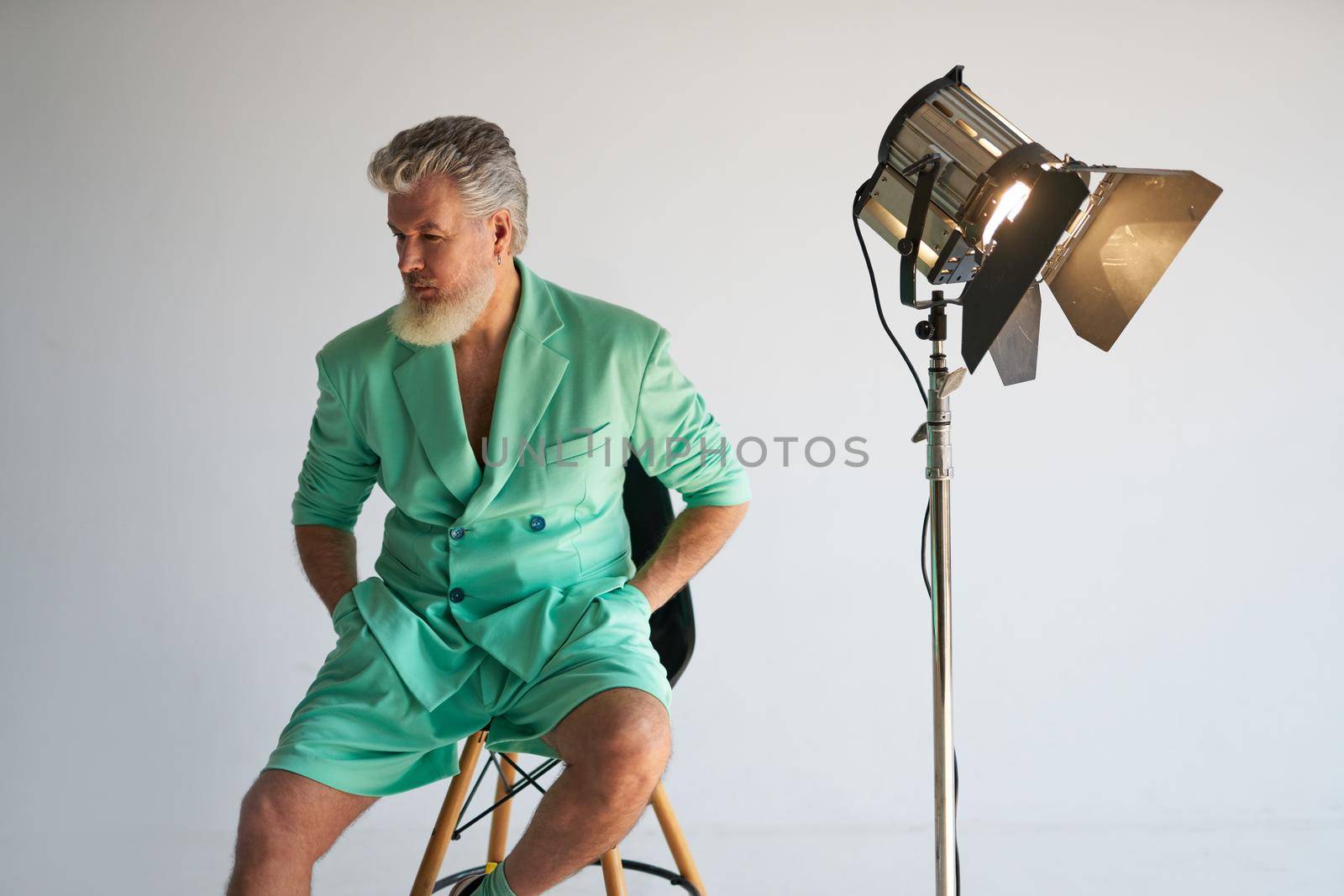 Studio shot of cool gray haired mature man wearing stylish outfit looking aside while sitting next to studio spotlight, posing over white background. Fashion photoshoot, style concept