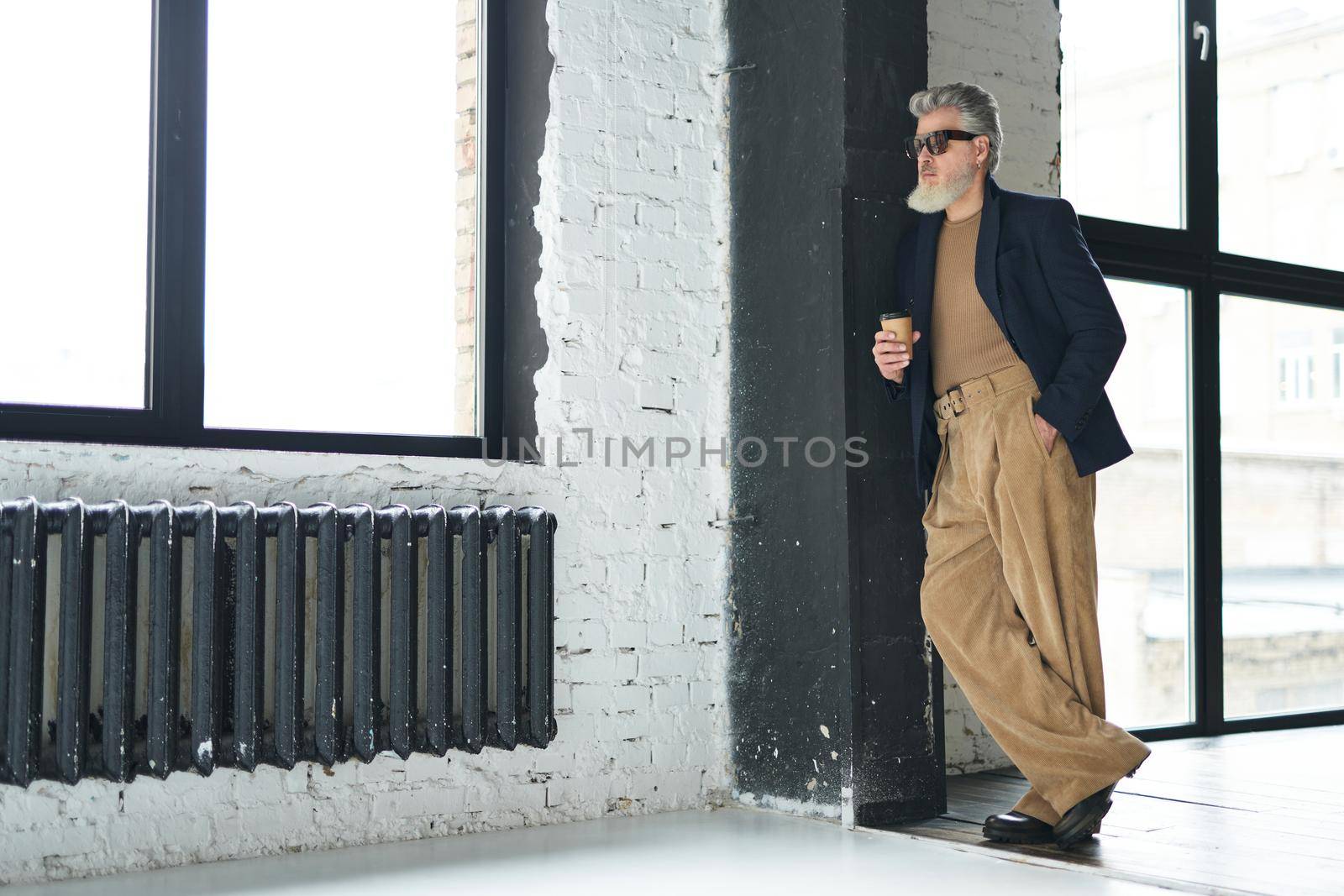 Full length shot of stylish middle aged man in business casual wear and sunglasses leaning on the wall, holding disposable coffee cup while posing in loft interior by friendsstock